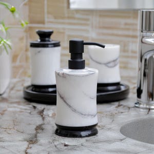 May include: A marble-patterned soap dispenser with a black pump, set on a countertop. The dispenser is white with gray veining and a black base. Other matching bathroom accessories are in the background, including a tumbler and a jar with a lid.