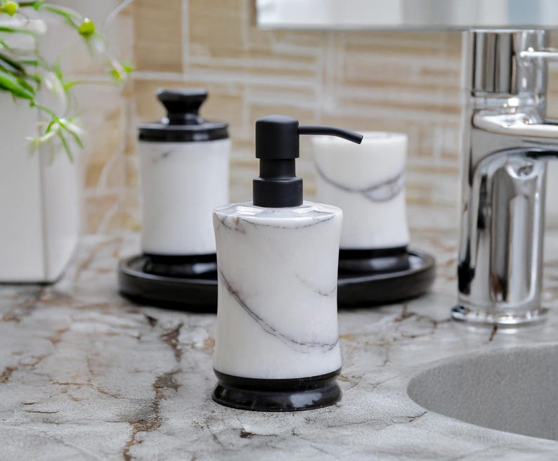 May include: A marble-patterned soap dispenser with a black pump, set on a countertop. The dispenser is white with gray veining and a black base. Other matching bathroom accessories are in the background, including a tumbler and a jar with a lid.