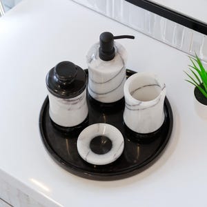 May include: A black marble bathroom set with white veining. The set includes a soap dispenser, a jar with a lid, a tumbler, and a small dish, all arranged on a black tray. A small potted plant is also present.