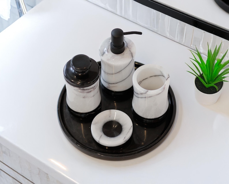 May include: A black marble bathroom set with white veining. The set includes a soap dispenser, a jar with a lid, a tumbler, and a small dish, all arranged on a black tray. A small potted plant is also present.