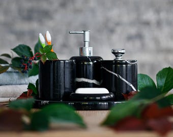 Può includere: Un set da bagno in marmo nero con venature bianche. Il set comprende un dosatore di sapone, un barattolo con coperchio, due portaspazzolini e un portasapone con una saponetta. Il set è esposto su un vassoio.
