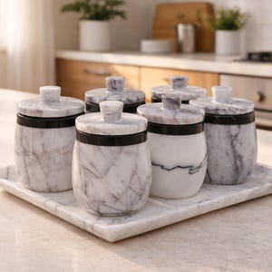May include: A set of six marble canisters with lids, each with a black band around the middle. The canisters are arranged on a matching marble tray. The marble has a gray and white veined pattern.
