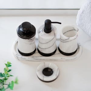 May include: A marble bathroom set featuring a soap dispenser, a jar with a lid, a small cup, and a soap dish. The set has black accents and a white and gray veined pattern. The items are arranged on a matching tray.