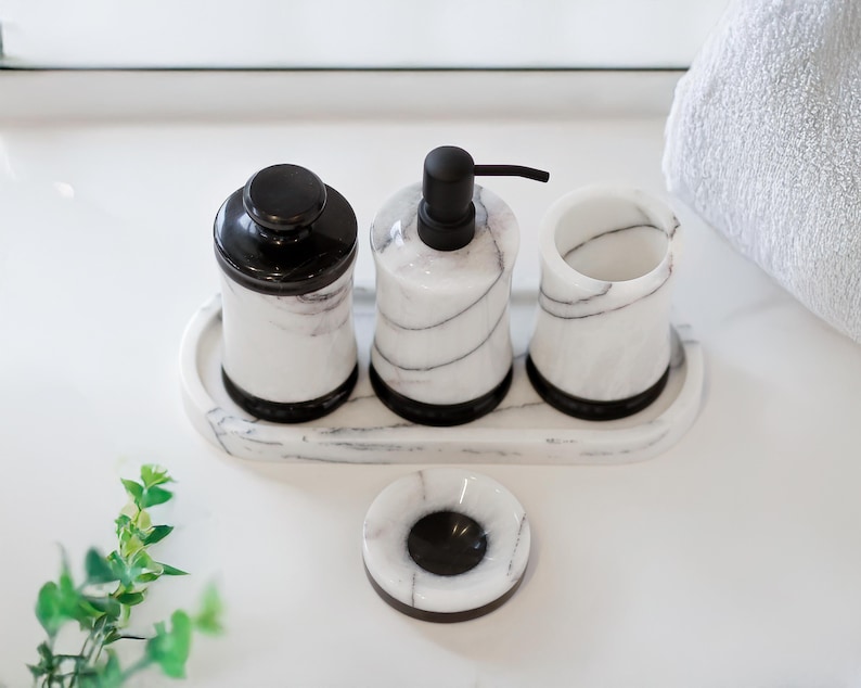 May include: A marble bathroom set featuring a soap dispenser, a jar with a lid, a small cup, and a soap dish. The set has black accents and a white and gray veined pattern. The items are arranged on a matching tray.