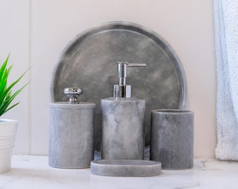May include: A gray marble bathroom set includes a soap dispenser, tumbler, soap dish, and a lidded jar. A matching gray marble tray sits behind the set. The set is on a white surface.