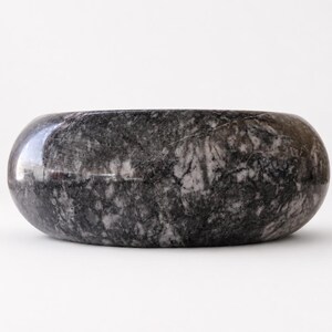May include: A circular bowl crafted from polished dark gray and black marble, featuring a smooth, rounded exterior surface with natural white veining patterns throughout the stone, presenting a solid, heavy appearance with a wide, open top.