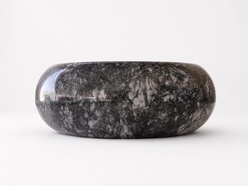 May include: A circular bowl crafted from polished dark gray and black marble, featuring a smooth, rounded exterior surface with natural white veining patterns throughout the stone, presenting a solid, heavy appearance with a wide, open top.