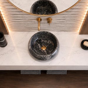 May include: A round black marble vessel sink sits on a white marble countertop with gray veining. The sink features a gold drain. A matching black marble tumbler and a black soap dish with a beige soap bar are placed on the countertop surface.