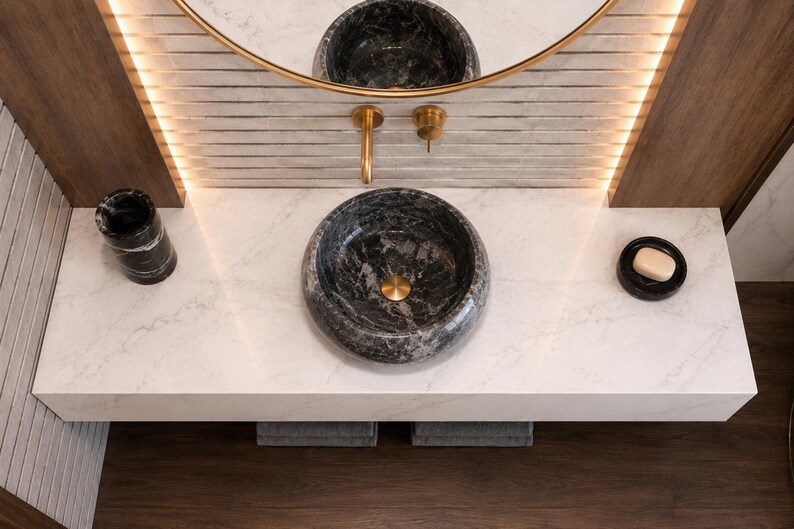 May include: A round black marble vessel sink sits on a white marble countertop with gray veining. The sink features a gold drain. A matching black marble tumbler and a black soap dish with a beige soap bar are placed on the countertop surface.