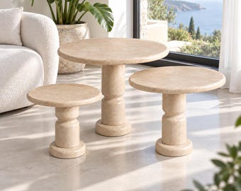 Op de afbeelding: Een set van drie ronde tafels met een beige, getextureerde afwerking die op steen lijkt. Elke tafel heeft een dikke, cilindrische voet met een inkeping in het midden en een breed, cirkelvormig tafelblad op verschillende hoogtes.