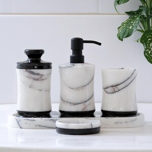 May include: A four-piece bathroom set made of white marble with black veining. The set includes a soap dispenser, a tumbler, a jar with a lid, and a soap dish. The dispenser has a black pump, and the other pieces have black accents.