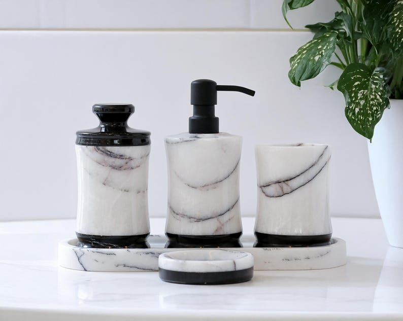 May include: A four-piece bathroom set made of white marble with black veining. The set includes a soap dispenser, a tumbler, a jar with a lid, and a soap dish. The dispenser has a black pump, and the other pieces have black accents.