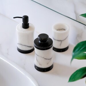 May include: A set of three bathroom accessories: a soap dispenser with a black pump, a jar with a black lid, and a tumbler. All are white with gray veining and black bases, arranged on a white marble surface.