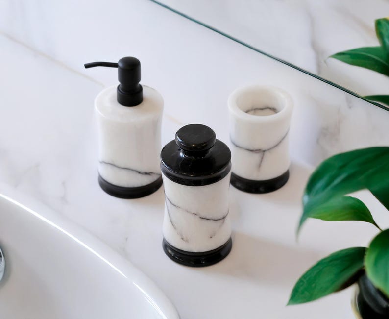 May include: A set of three bathroom accessories: a soap dispenser with a black pump, a jar with a black lid, and a tumbler. All are white with gray veining and black bases, arranged on a white marble surface.