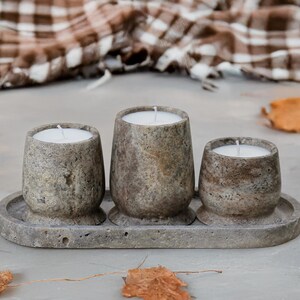 Puede incluir: Tres candelabros de piedra con velas blancas sobre una bandeja a juego. Los candelabros son de diferentes tamaños y tienen un aspecto texturizado y moteado. El fondo incluye una manta a cuadros y hojas caídas.