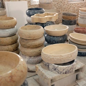 May include: A collection of stone vessel sinks in various shapes, sizes, and colors including beige, gray, white, and dark blue. The sinks are stacked and arranged in a workshop, featuring smooth, polished, and textured natural stone finishes.