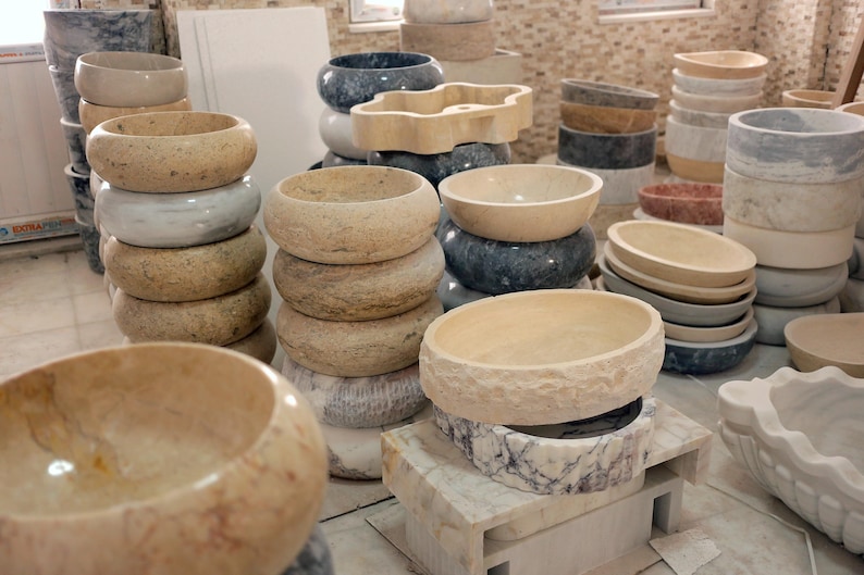 May include: A collection of stone vessel sinks in various shapes, sizes, and colors including beige, gray, white, and dark blue. The sinks are stacked and arranged in a workshop, featuring smooth, polished, and textured natural stone finishes.