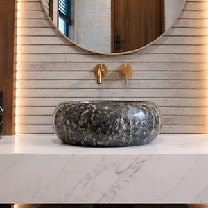 May include: A round, dark gray and black speckled stone vessel sink sits on a white marble countertop. Above the sink, a gold wall-mounted faucet is centered against a light gray horizontal slatted wall with vertical LED accent lighting.