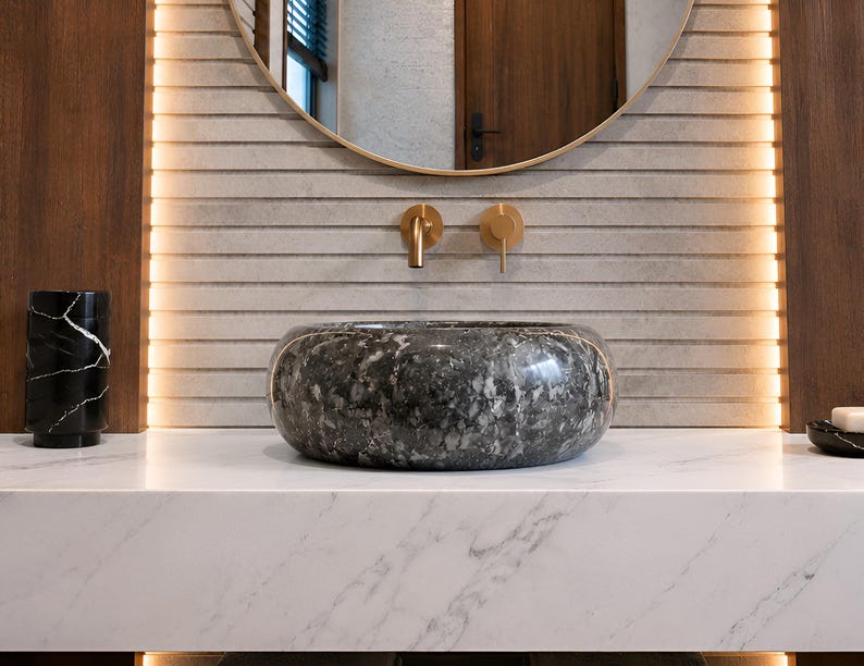 May include: A round, dark gray and black speckled stone vessel sink sits on a white marble countertop. Above the sink, a gold wall-mounted faucet is centered against a light gray horizontal slatted wall with vertical LED accent lighting.