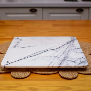 May include: A white marble serving board with gray veining, sitting on a woven rope mat on a wooden table.