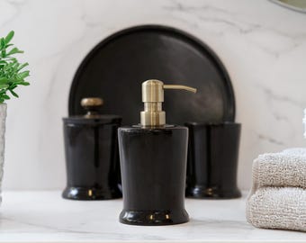 Può includere: Un dispenser di sapone in ceramica nera con una pompa color oro è il punto focale di questo set da bagno. Il set include un bicchiere abbinato, un barattolo con coperchio e un vassoio nero. Gli articoli sono disposti su una superficie di marmo bianco.