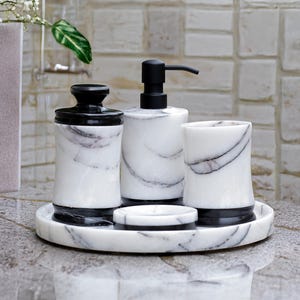 May include: A marble bathroom set featuring a soap dispenser, tumbler, jar, and soap dish. The set has a black and white color scheme with a tray to hold the items. The items have a black base and lid, with a white marble design.