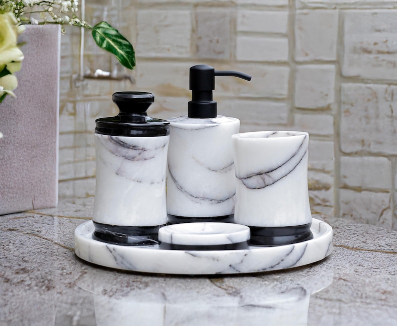 May include: A marble bathroom set featuring a soap dispenser, tumbler, jar, and soap dish. The set has a black and white color scheme with a tray to hold the items. The items have a black base and lid, with a white marble design.