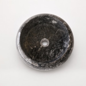 May include: A circular vessel sink crafted from dark gray and black natural stone with a polished finish. The basin features a smooth, rounded rim and a centered circular drain hole, showcasing the unique veining and mottled texture of the stone.