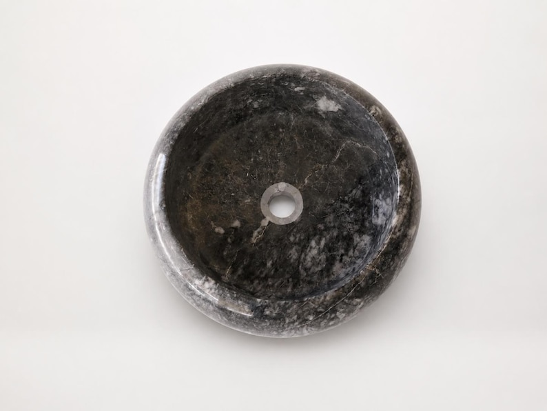 May include: A circular vessel sink crafted from dark gray and black natural stone with a polished finish. The basin features a smooth, rounded rim and a centered circular drain hole, showcasing the unique veining and mottled texture of the stone.