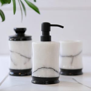 May include: A white marble soap dispenser with a black pump, a matching jar with a black lid, and a tumbler. The bathroom accessories have black bases and veining throughout the white marble.