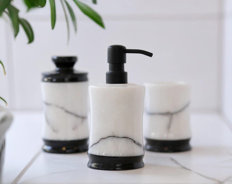 May include: A white marble soap dispenser with a black pump, a matching jar with a black lid, and a tumbler. The bathroom accessories have black bases and veining throughout the white marble.