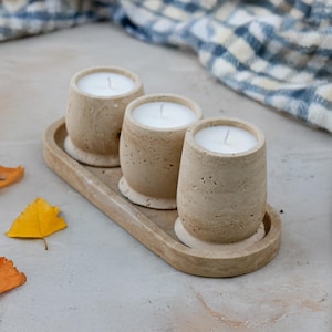 Puede incluir: Tres velas de piedra beige con cera blanca, colocadas sobre una bandeja ovalada a juego. Las velas son cilíndricas con base redondeada. Hojas de otoño y un paño a cuadros azul y blanco en el fondo.