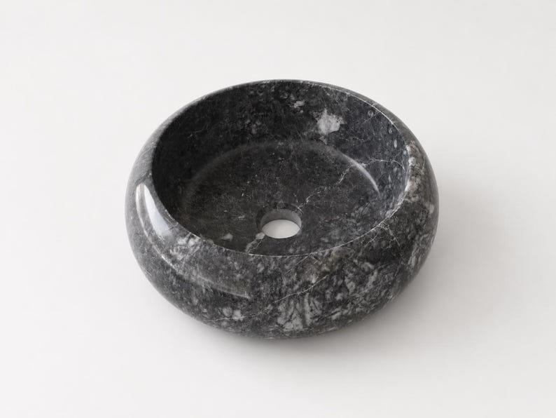 May include: A round vessel sink crafted from dark gray and black marble with prominent white veining throughout the stone. The basin features a smooth, polished finish and a centered circular drain hole at the bottom of the bowl interior.