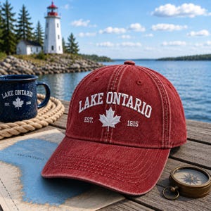 May include: A red denim baseball cap featuring white embroidered text that reads LAKE ONTARIO and EST. 1615, centered with a white maple leaf icon. The cap has a structured crown, a curved brim, and visible stitching details across the fabric panels.