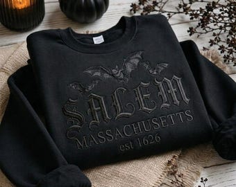 May include: A black long-sleeve sweatshirt featuring black embroidery of three bats above the text SALEM MASSACHUSETTS est 1626 in a gothic font. The garment has a crew neckline and a ribbed collar, cuffs, and hem, showcasing a monochromatic design.