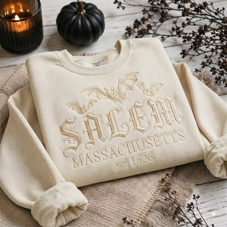 May include: A cream-colored crewneck sweatshirt featuring embroidered text that reads SALEM MASSACHUSETTS est 1626. The design includes stitched bat illustrations above the lettering, all rendered in a matching light tan thread color.