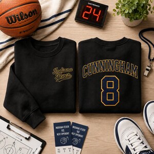 Op de afbeelding: Twee zwarte sweaters met ronde hals. E&eacute;n heeft de gouden tekst Indiana Fever op de borst. De andere toont de gouden tekst CUNNINGHAM boven het grote goud-blauwe nummer 8 op de achterkant. De items zijn gerangschikt met een basketbal, een digitaal scorebord met 24, twee wedstrijdkaartjes, een klembord en een sneaker.