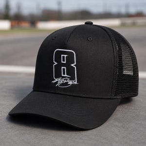 Może przedstawiać: Czarna czapka typu trucker z białym numerem 8 i podpisem. Czapka ma siatkowy tył i zakrzywiony daszek. Wykonana z wytrzymałego materiału.