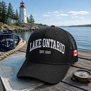 Op de afbeelding: Een zwarte truckerpet met witte geborduurde tekst die LAKE ONTARIO EST. 1615 leest op het voorpaneel. De pet bevat een kleine Canadese vlag-patch aan de zijkant en een achterpaneel van gaas voor ventilatie, ontworpen met een gestructureerde kroon.