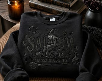 May include: A black crewneck sweatshirt featuring an embroidered graphic of a witch in a hat sitting on a stool. The design includes the text "SALEM" and "MASSACHUSETTS" along with illustrations of a crescent moon, stars, a mortar and pestle, potion bottles, and a broomstick.