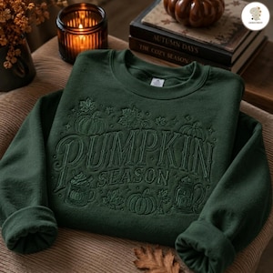 Peut inclure: Un sweat-shirt à col rond vert forêt foncé avec une broderie ton sur ton du texte PUMPKIN SEASON. Le motif comprend des illustrations cousues de citrouilles, de feuilles d'érable, d'une tasse avec de la crème fouettée et d'une lanterne.