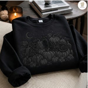 Puede incluir: Una sudadera negra de cuello redondo con un diseño bordado en tonos oscuros que muestra tres gatos sentados entre varias calabazas. La escena incluye una luna creciente, pequeñas estrellas y detalles de enredaderas en un estilo monocromático.