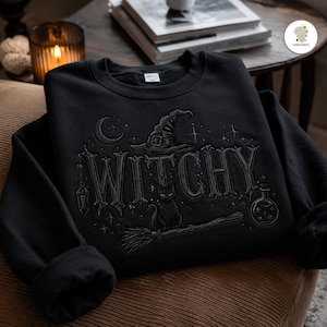 Peut inclure: Un sweat-shirt noir à col rond avec un motif brodé représentant le mot WITCHY en grandes lettres gothiques. Le graphisme comprend un chapeau de sorcière, un croissant de lune, des étoiles, un chat noir, un balai, une lanterne et une fiole.