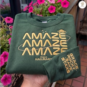 May include: A dark green sweatshirt with the word "AMAZE" stacked three times in gold lettering, with a turtle graphic and the words "Project Hailmary". The cuff displays "GRACE ROCKY SAVE STARS" in gold.