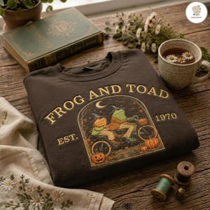 Könnte beinhalten: Ein dunkelbraunes Sweatshirt mit Rundhalsausschnitt und einer Stickerei von zwei Fröschen auf einem Tandemfahrrad. Der Text "FROG AND TOAD" und "EST. 1970" ist in Goldfaden gestickt. Das Motiv zeigt Kürbisse, Herbstblätter und eine Mondsichel.