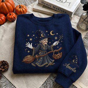 May include: A navy blue crewneck sweatshirt featuring an embroidered graphic of a witch in a pointed hat riding a broomstick, surrounded by gold stars and moons. The sleeve displays an embroidered witch hat above the name Amela in gold script.