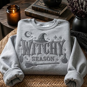 Peut inclure: Un sweat-shirt gris clair à col rond avec une broderie gris foncé indiquant "WITCHY SEASON" dans une police stylisée. Le motif comprend un chapeau de sorcière, un croissant de lune, des étoiles, un pendentif cristal, une fiole et un balai.