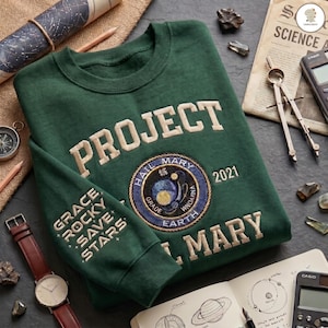 Peut inclure: Un sweat-shirt &agrave; manches longues vert fonc&eacute; avec le texte brod&eacute; PROJECT HAIL MARY et l'ann&eacute;e 2021. Un &eacute;cusson circulaire repr&eacute;sente une sc&egrave;ne spatiale avec le texte HAIL MARY GRADE EARTH. La manche porte le texte GRACE ROCKY SAVE STARS.