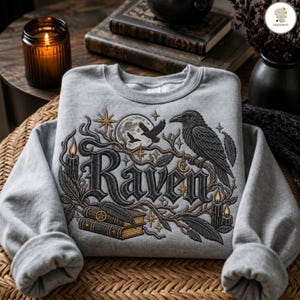 Könnte beinhalten: Ein graumeliertes Sweatshirt mit Rundhalsausschnitt und einer Stickerei, die einen großen Raben auf einem Ast, das Wort Raven in gotischer Schrift, zwei gestapelte Bücher mit Pentagramm, einen Vollmond, Sterne und Kerzen zeigt.