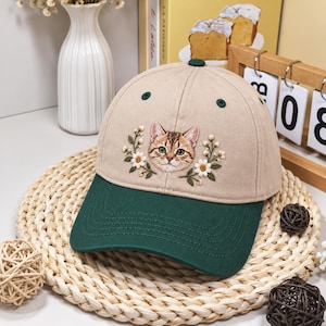 May include: A beige baseball cap with a dark green brim featuring an embroidered brown tabby cat face surrounded by white daisy flowers and green leaves. The cap has a small dark green button on the top and dark green stitched eyelets.